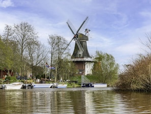 Eine traditionelle Windmühle an einem ruhigen Fluss, in dessen Nähe Boote liegen.