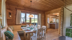 A wooden cabin interior with a dining area, a sofa, and a kitchen visible through an open doorway.