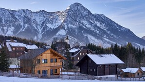 A wooden chalet with a snow-covered roof, surrounded by a wooden fence, and a mountain backdrop.