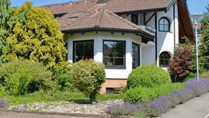 Ein zweistöckiges Haus mit Ziegeldach, großen Fenstern und einem Garten mit verschiedenen Pflanzen und Blumen.