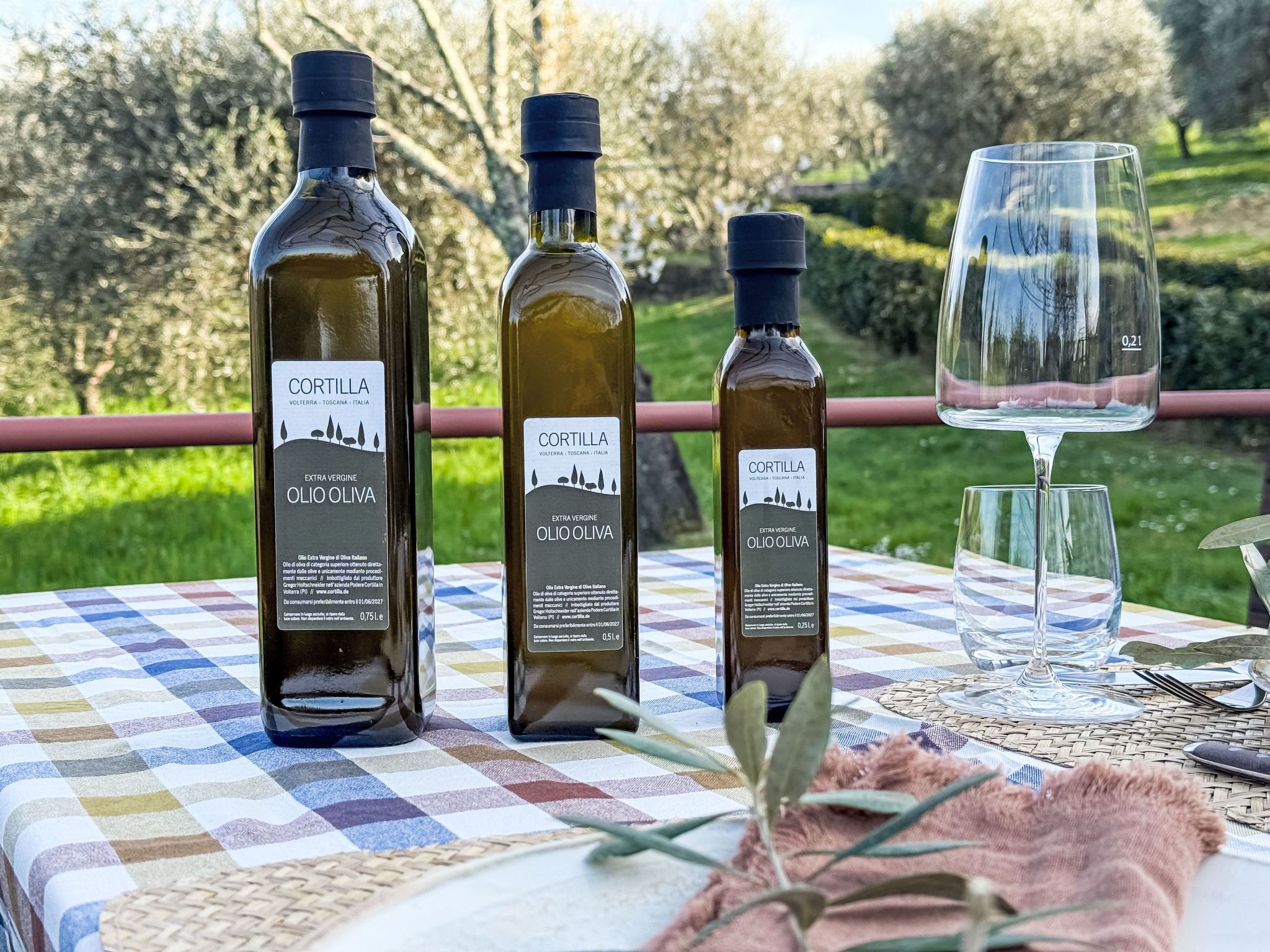 Three bottles of extra virgin olive oil and a wine glass on a table with a checkered tablecloth.