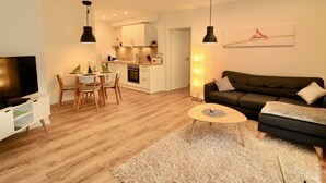 Ein modernes Wohnzimmer mit Flachbildfernseher, einem dunkelgrauen Sofa, einem runden Couchtisch und einem Essbereich mit kleinem Tisch und Stühlen.