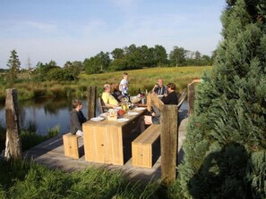Eine Holzterrasse mit gedecktem Tisch, umgeben von einer natürlichen Landschaft mit Bäumen und einem Gewässer.