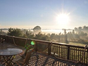 Eine Holzterrasse mit Tisch und Stühlen, die auf eine nebelverhangene Landschaft mit Bäumen und einem Gewässer blickt.