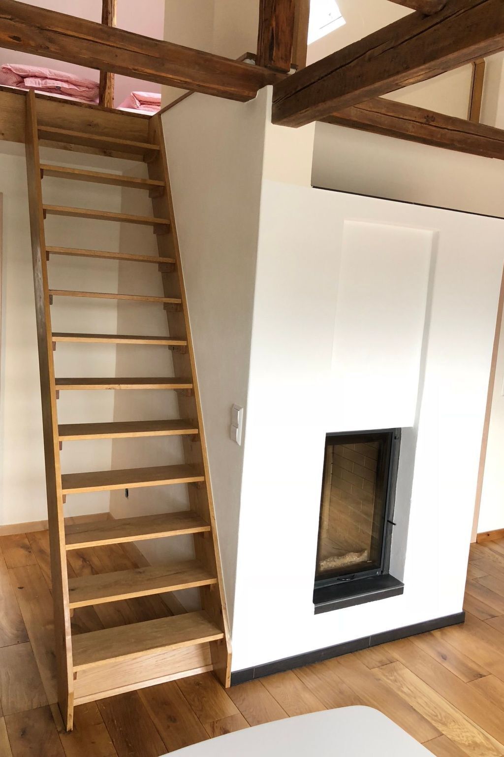 A room with a wooden ladder leading to a loft area, a white wall with a fireplace, and wooden flooring.