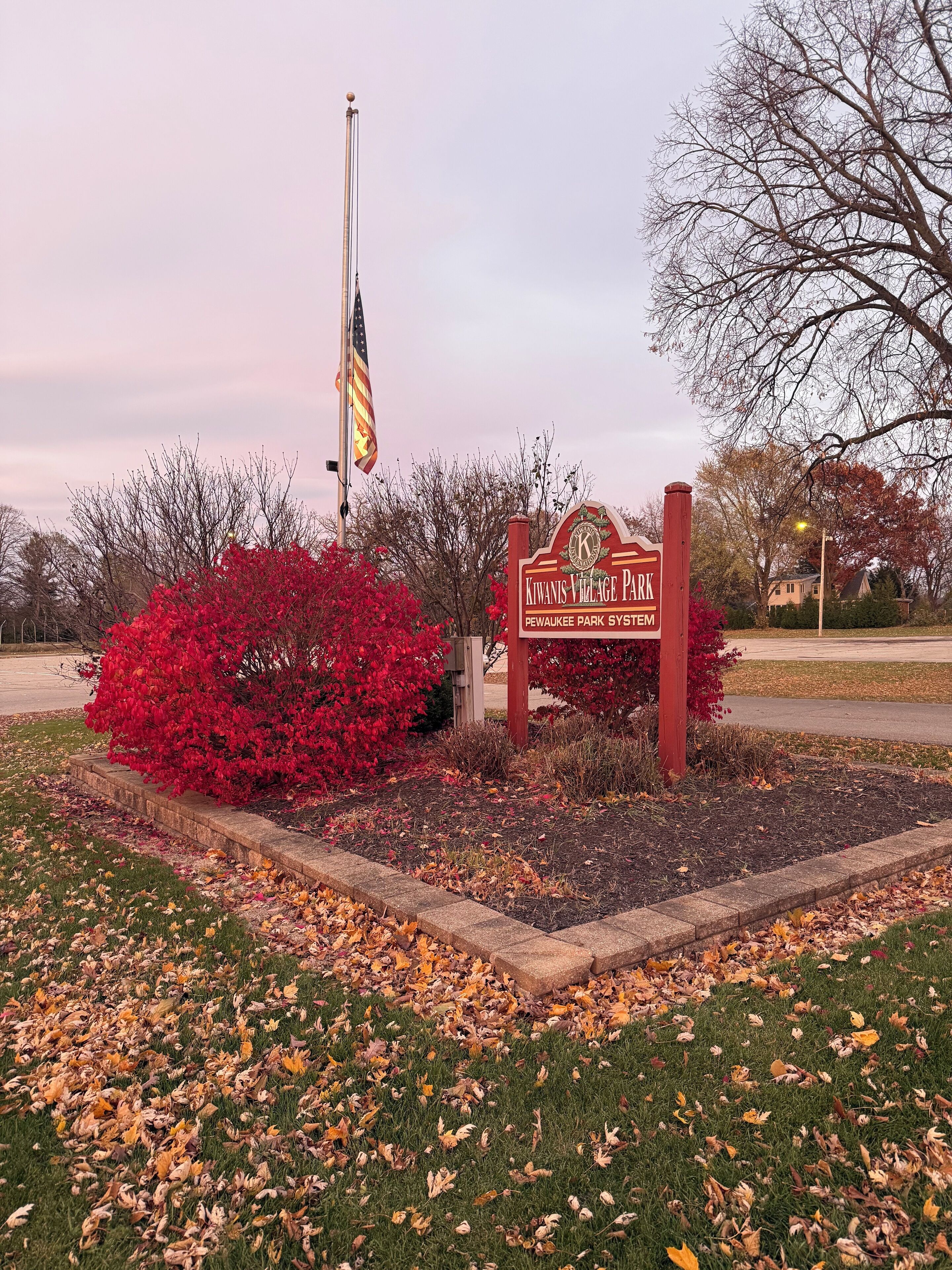 A sign for Kiwanis Village Park with a flagpole, red flowering bushes, and autumn leaves.