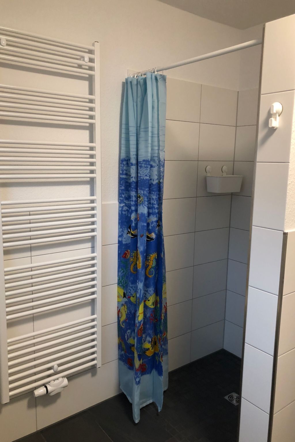 Ducha con cortina azul decorada con motivos de peces, toallero blanco y estante pequeño blanco.
