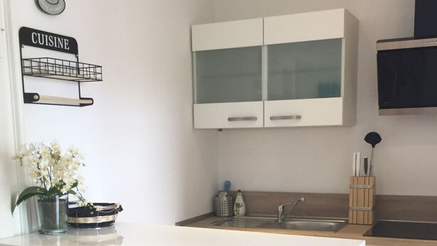 Eine moderne Küche mit weißen Schränken, einer Edelstahlspüle und einer weißen Arbeitsplatte. Auf der Arbeitsplatte stehen drei weiße Barhocker und eine Vase mit weißen Blumen.