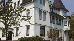 Ein zweistöckiges weißes Haus mit dunklem Dach, einer Veranda und einem kleinen Garten.