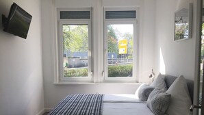 Ein Schlafzimmer mit einem Bett, zwei Fenstern, einem an der Wand befestigten Fernseher und einer Tür.