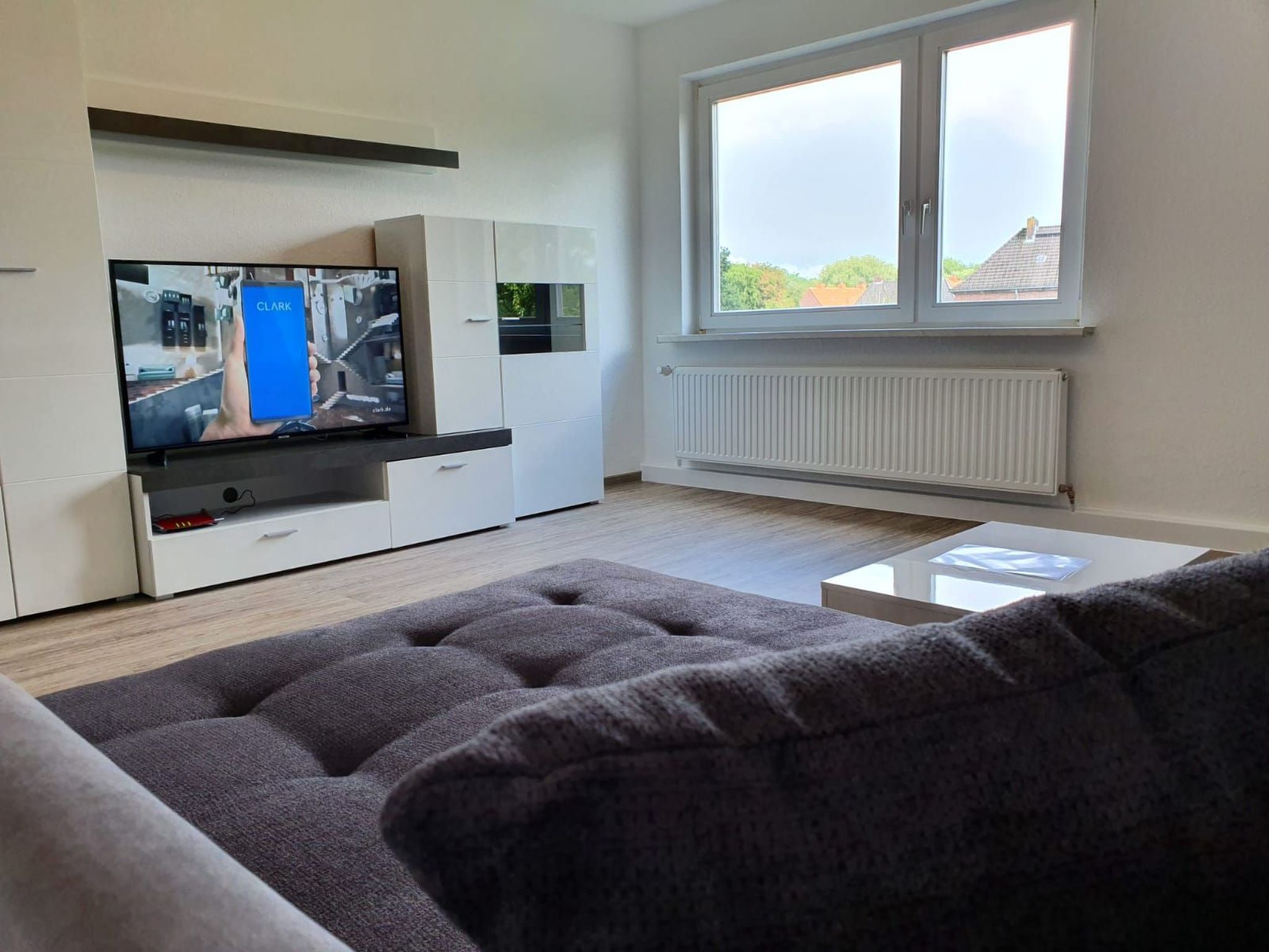 A modern living room with a flat-screen TV, white cabinetry, a grey sofa, and a window with a view of houses.