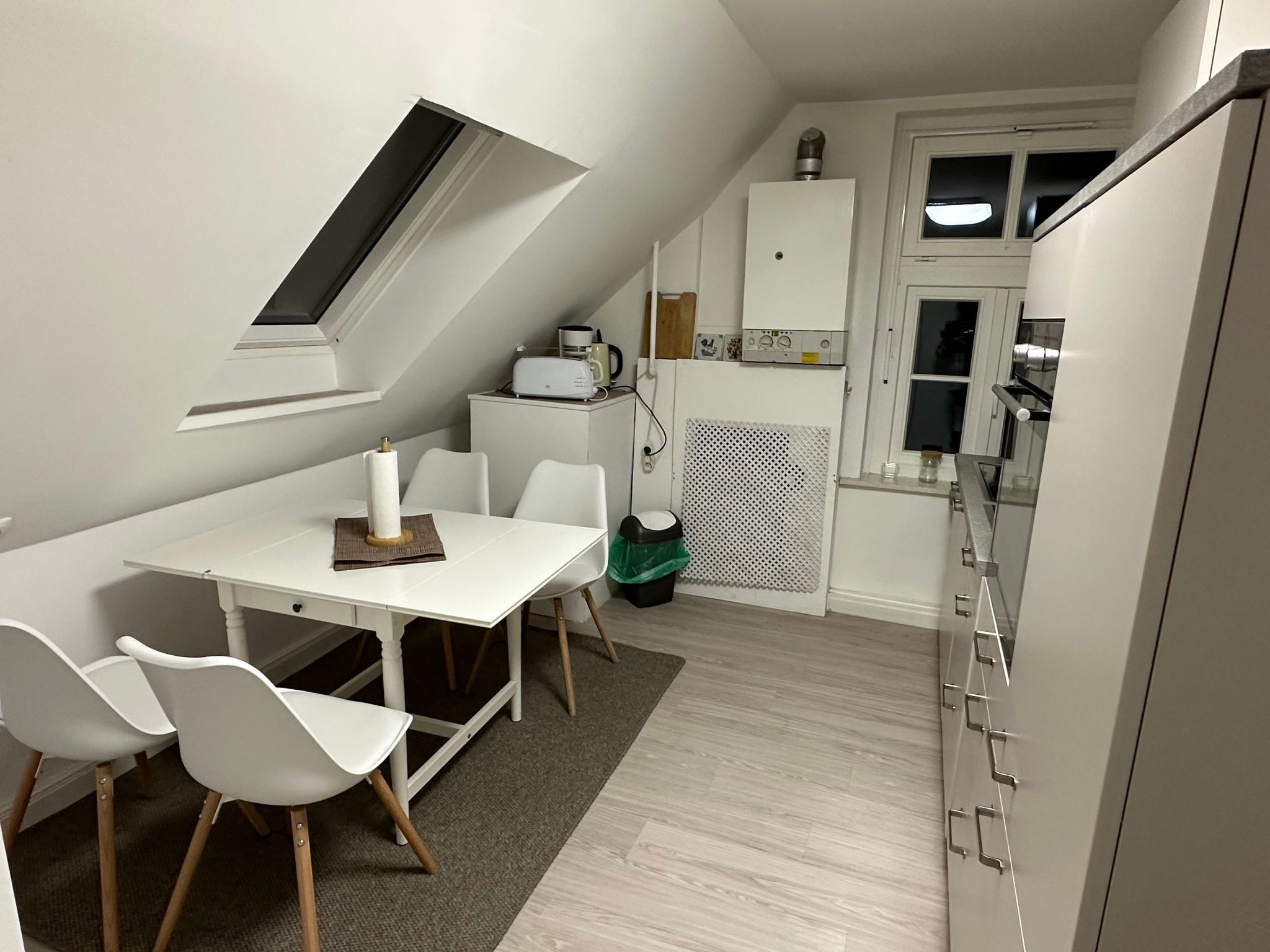 Eine kompakte Küche mit weißen Schränken, einem eingebauten Backofen und einer Mikrowelle. Es gibt einen Essbereich mit einem weißen Tisch und Stühlen, sowie ein kleines Fenster über der Spüle.