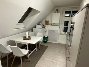 Eine kompakte Küche mit weißen Schränken, einem eingebauten Backofen und einer Mikrowelle. Es gibt einen Essbereich mit einem weißen Tisch und Stühlen, sowie ein kleines Fenster über der Spüle.