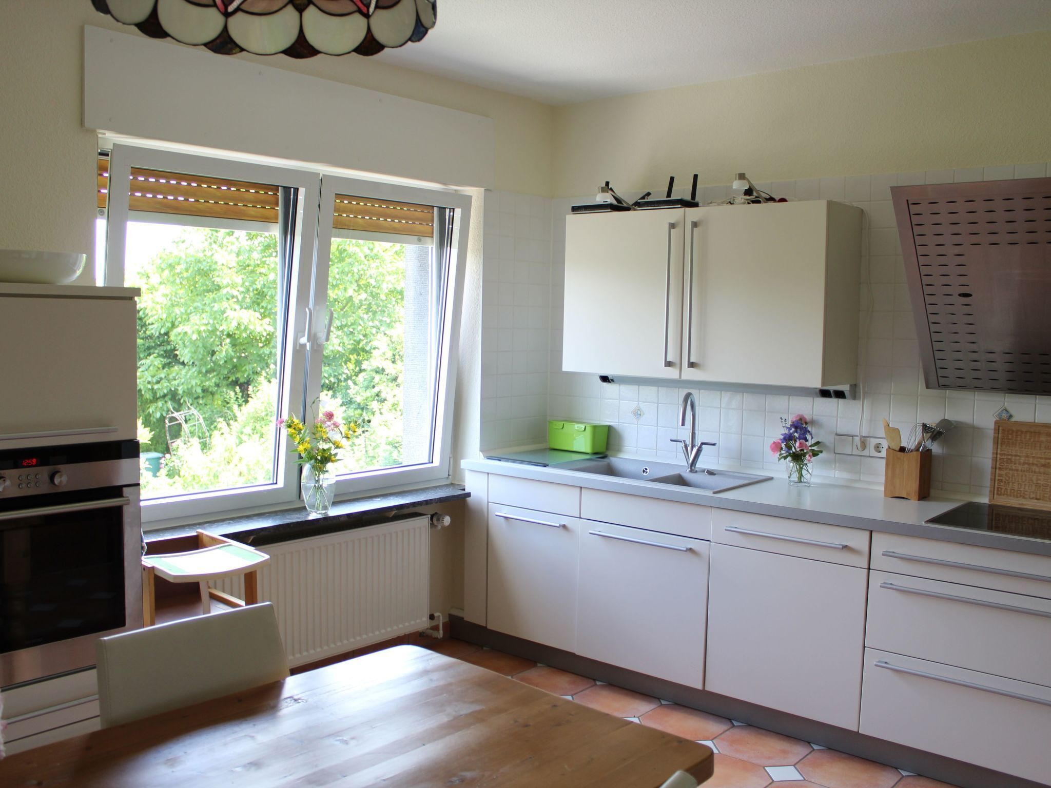 Une cuisine avec des meubles blancs, une table en bois, une fenêtre avec des stores et un radiateur.