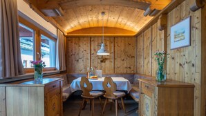 Ein Holzinterieur mit einem Essbereich, der einen Tisch und Stühle, ein Fenster mit Vorhängen sowie eine Holzkommode beinhaltet.