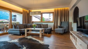 Ein modernes Wohnzimmer mit Sofa, Hocker, Couchtisch und Fernseher. Ein großes Fenster mit Vorhängen, Holzboden und ein Kuhfellteppich sind ebenfalls zu sehen.