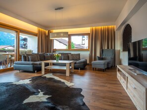 Ein modernes Wohnzimmer mit Sofa, Hocker, Couchtisch und Fernseher. Ein großes Fenster mit Vorhängen, Holzboden und ein Kuhfellteppich sind ebenfalls zu sehen.