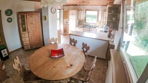 A wooden dining table with chairs, a kitchen area with wooden cabinets, a window with a view of greenery, and a ceiling with wooden beams.