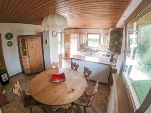 Una mesa de comedor de madera con sillas, una zona de cocina con armarios de madera, una ventana con vistas a la vegetación y un techo con vigas de madera.