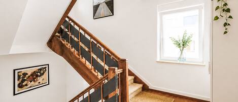 A staircase with wooden railings and a black handrail, leading to a landing with a framed picture and a window with a view of greenery.