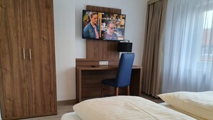 Ein Hotelzimmer mit einem Holzschrank, einem Schreibtisch mit Stuhl, einem über dem Schreibtisch angebrachten Fernseher und einem Bett mit weißer Daunendecke.