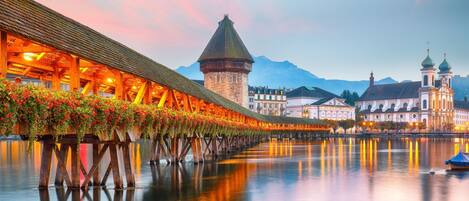 A wooden bridge with flower boxes, a historic building with domes, and a calm river reflecting the structures.