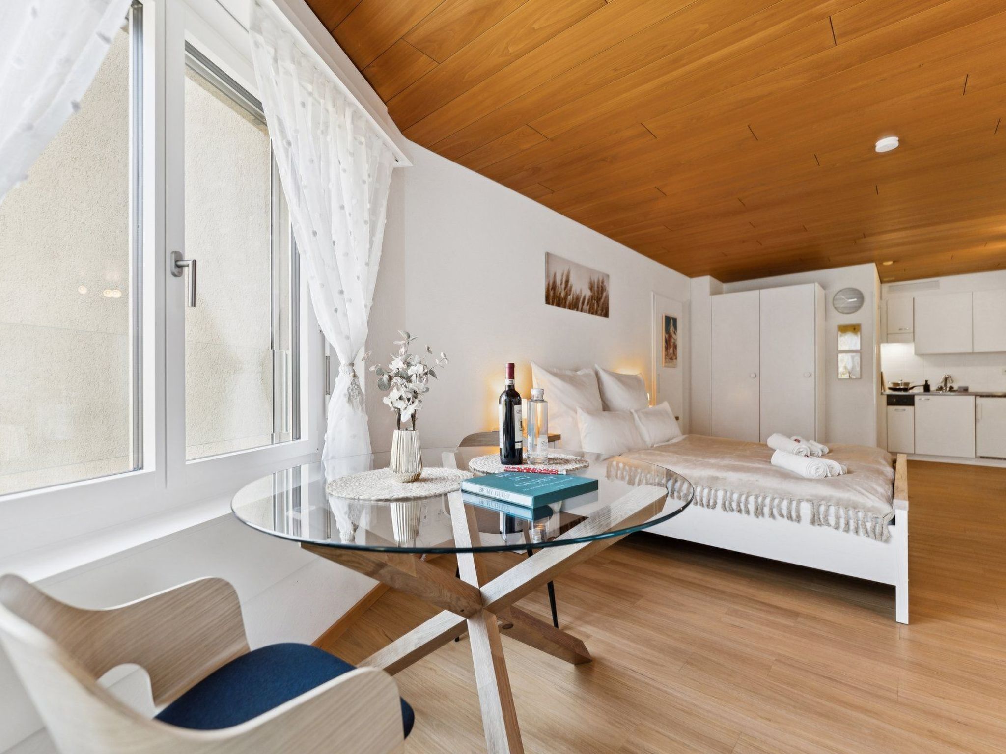 A modern bedroom with a glass table, a bed with white bedding, a small round table with a vase and bottles, and a view of a kitchen area.