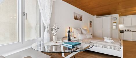A modern bedroom with a glass table, a bed with white bedding, a small round table with a vase and bottles, and a view of a kitchen area.