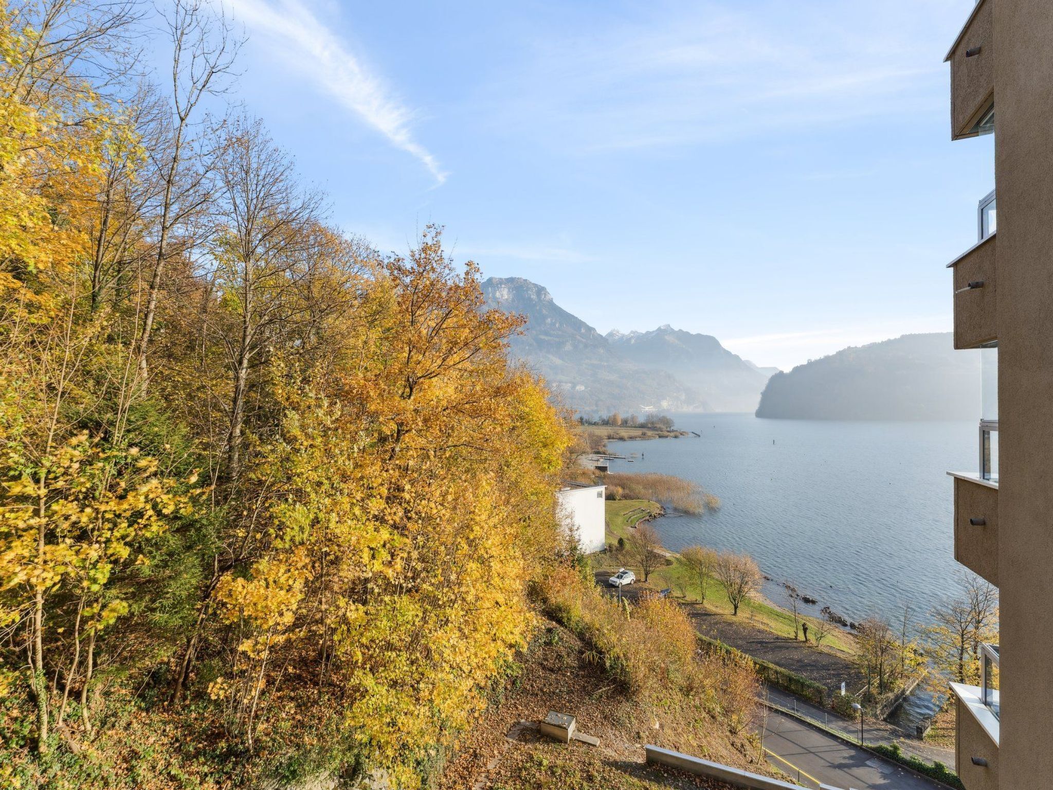A view of a lake with autumn foliage, a building with a balcony overlooking the water, and distant mountains.
