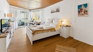Quarto com teto de madeira, cama com roupa de cama branca, mesa de cabeceira com abajur, janela com cortinas e um pôster na parede.