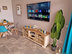 Ein modernes Wohnzimmer mit einem Flachbildfernseher, der Streaming-Apps anzeigt, einem Holzkonsolentisch mit Dekoration, einer Essecke mit Stühlen und einem Tisch sowie einer großen Zimmerpflanze.
