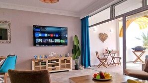 A modern living room with a flat-screen TV, a wooden cabinet with decorative letters, a dining area with a fruit bowl, and a view through a glass door. - Vista Faro by Interhome (Morro Jable)