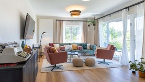 A modern living room with a sofa, armchairs, a coffee table, and a television. There are plants, a lamp, and a large window with curtains. - Overlook Lodge by Interhome (Vöhl)