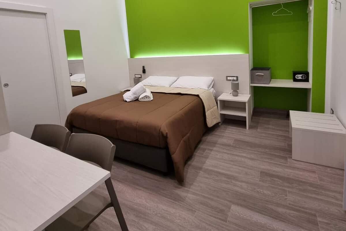A modern hotel room with a large bed, a desk, a chair, a wardrobe, and a mirror.