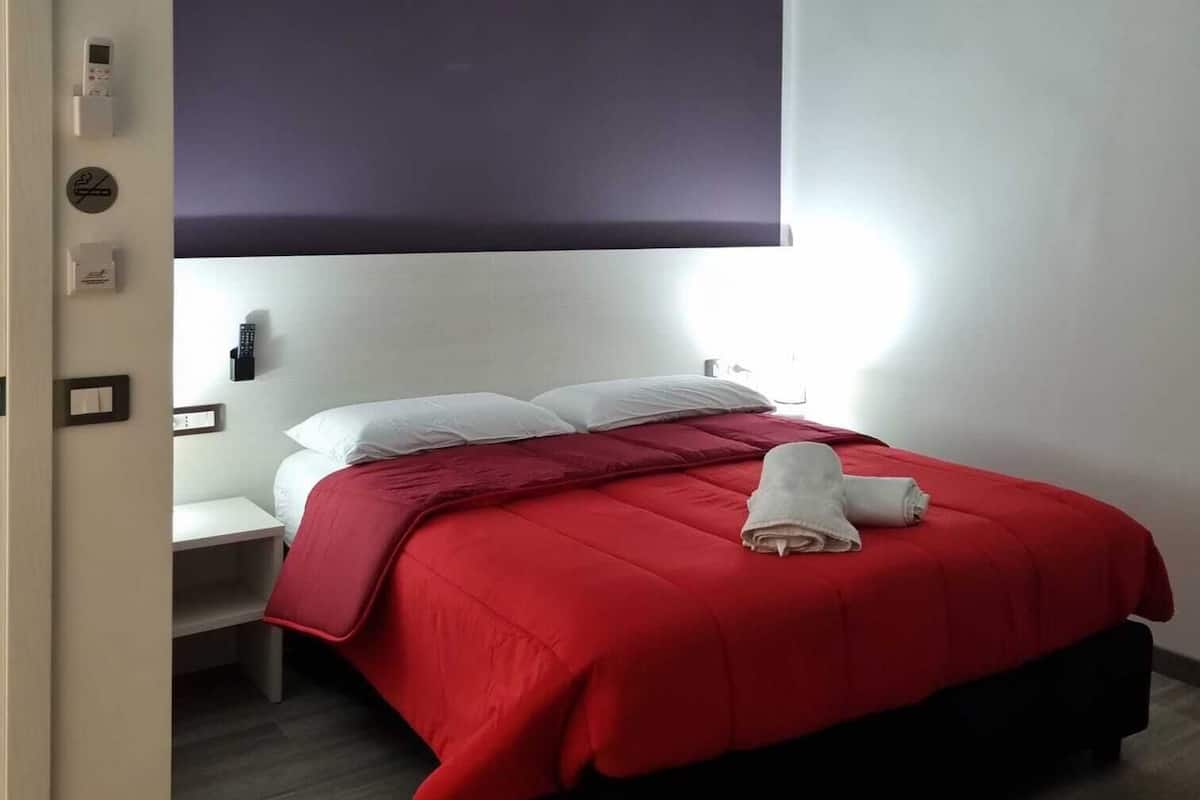 A hotel room with a bed, a nightstand, a wall-mounted control panel, and a door.