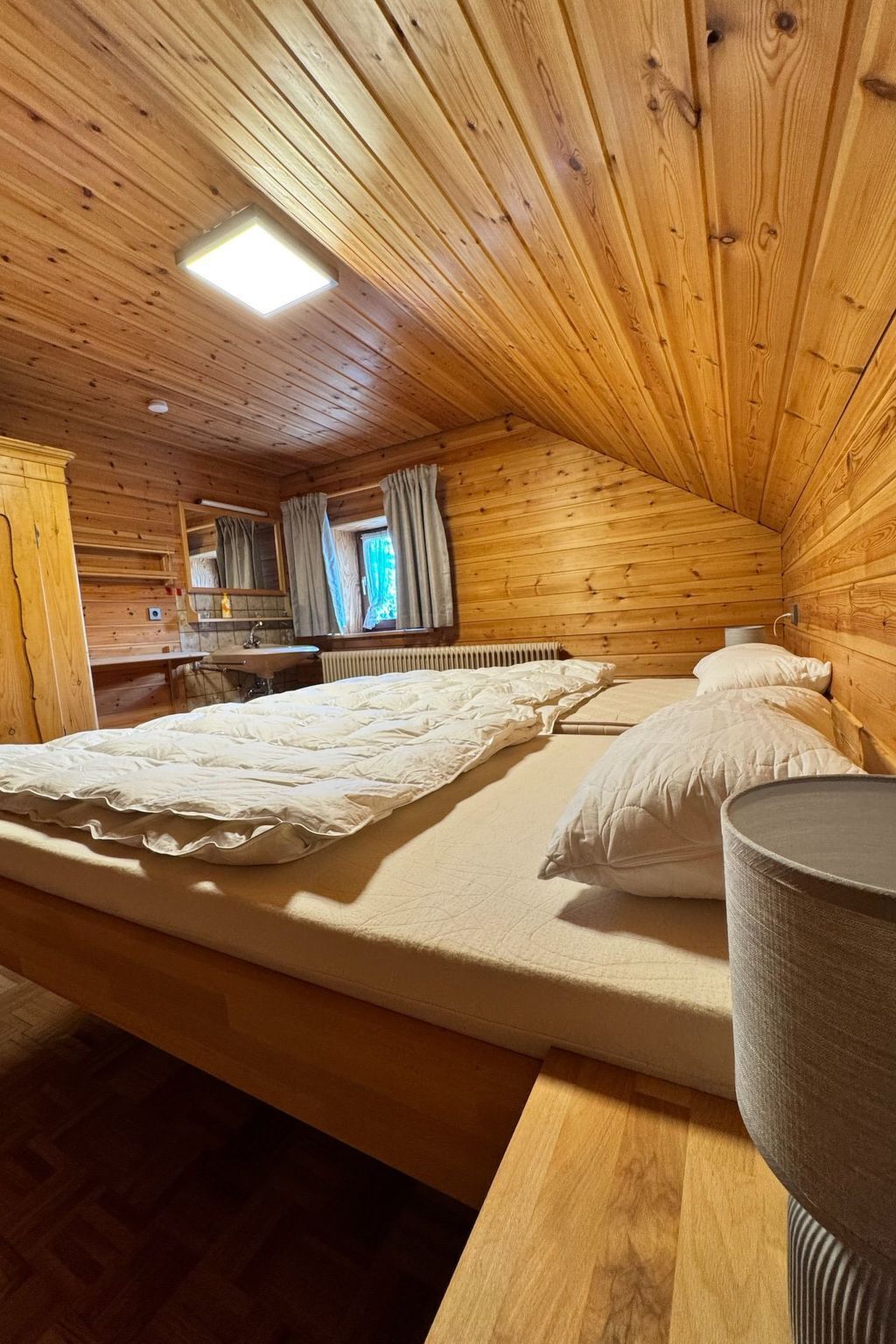 A wooden cabin room with a bed, a small table, a window with curtains, and a radiator.