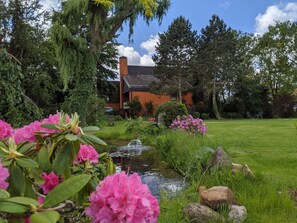 Ein Haus mit einem Ziegelkamin, umgeben von einem üppigen Garten mit einem kleinen Teich und rosa Blumen.