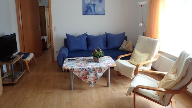 Een woonkamer met een blauwe bank, een witte fauteuil, een kleine tafel met een bloemrijk tafelkleed en een televisie op een dressoir.