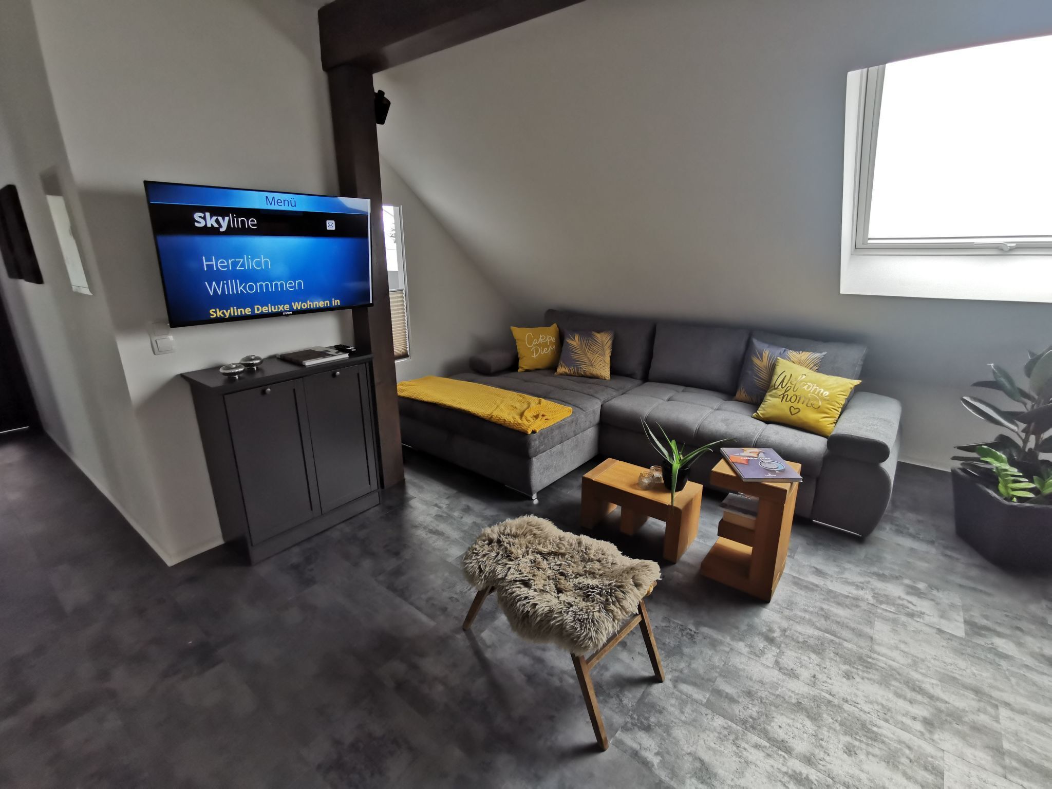 A modern living room with a flat-screen TV, grey sofa with yellow cushions, a small wooden coffee table, and a fluffy chair.
