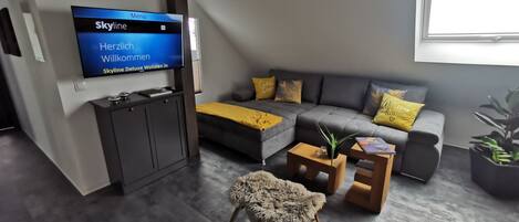 A modern living room with a flat-screen TV, grey sofa with yellow cushions, a small wooden coffee table, and a fluffy chair.