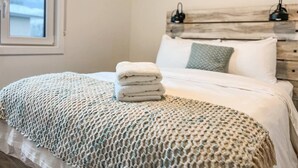 Un lit bien fait, avec une couverture à motifs et des serviettes pliées, une tête de lit en bois et un tableau accroché au-dessus.