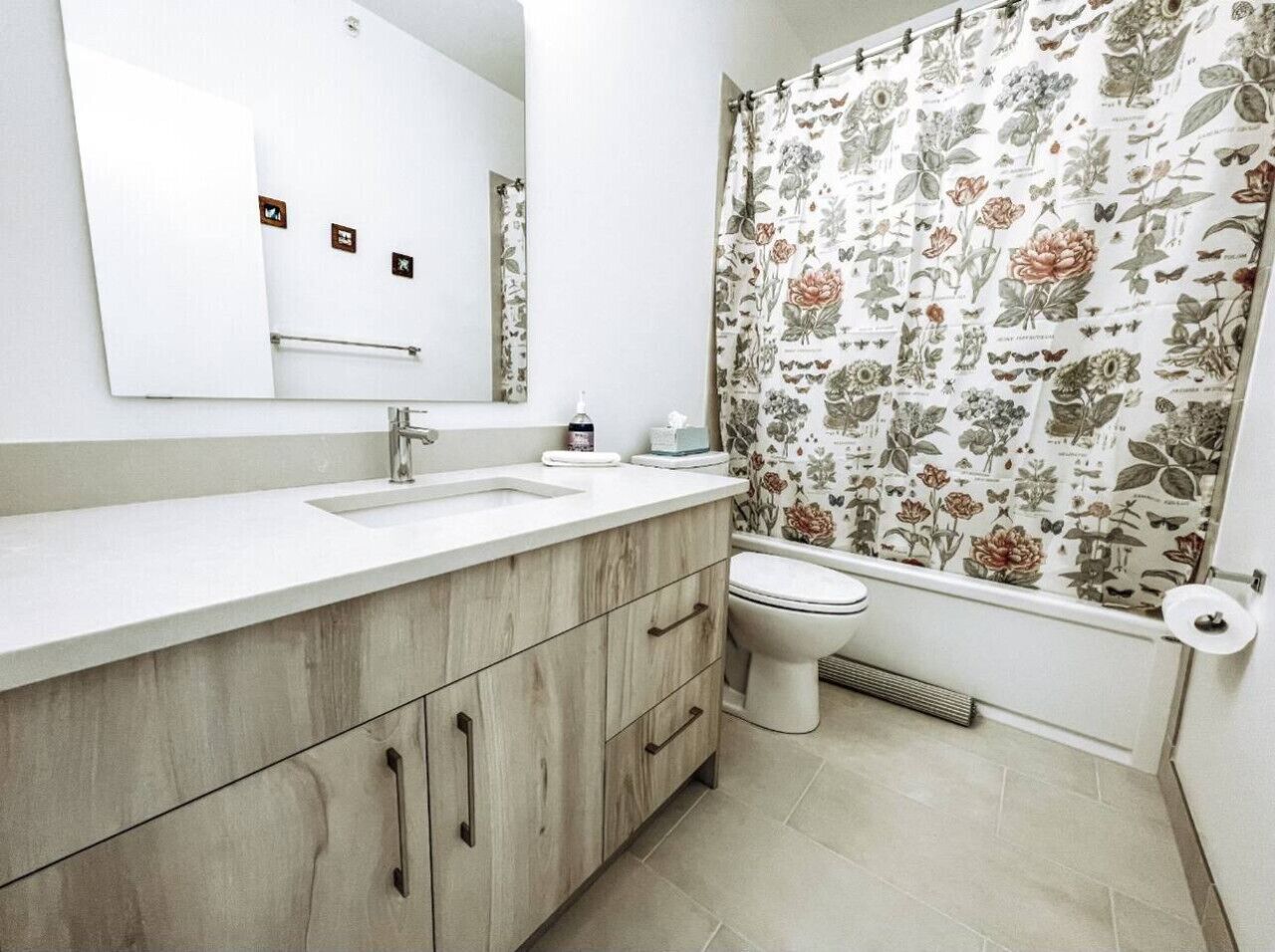 Un baño con lavamanos blanco, armarios de madera, inodoro y cortina de ducha estampada con motivos florales.