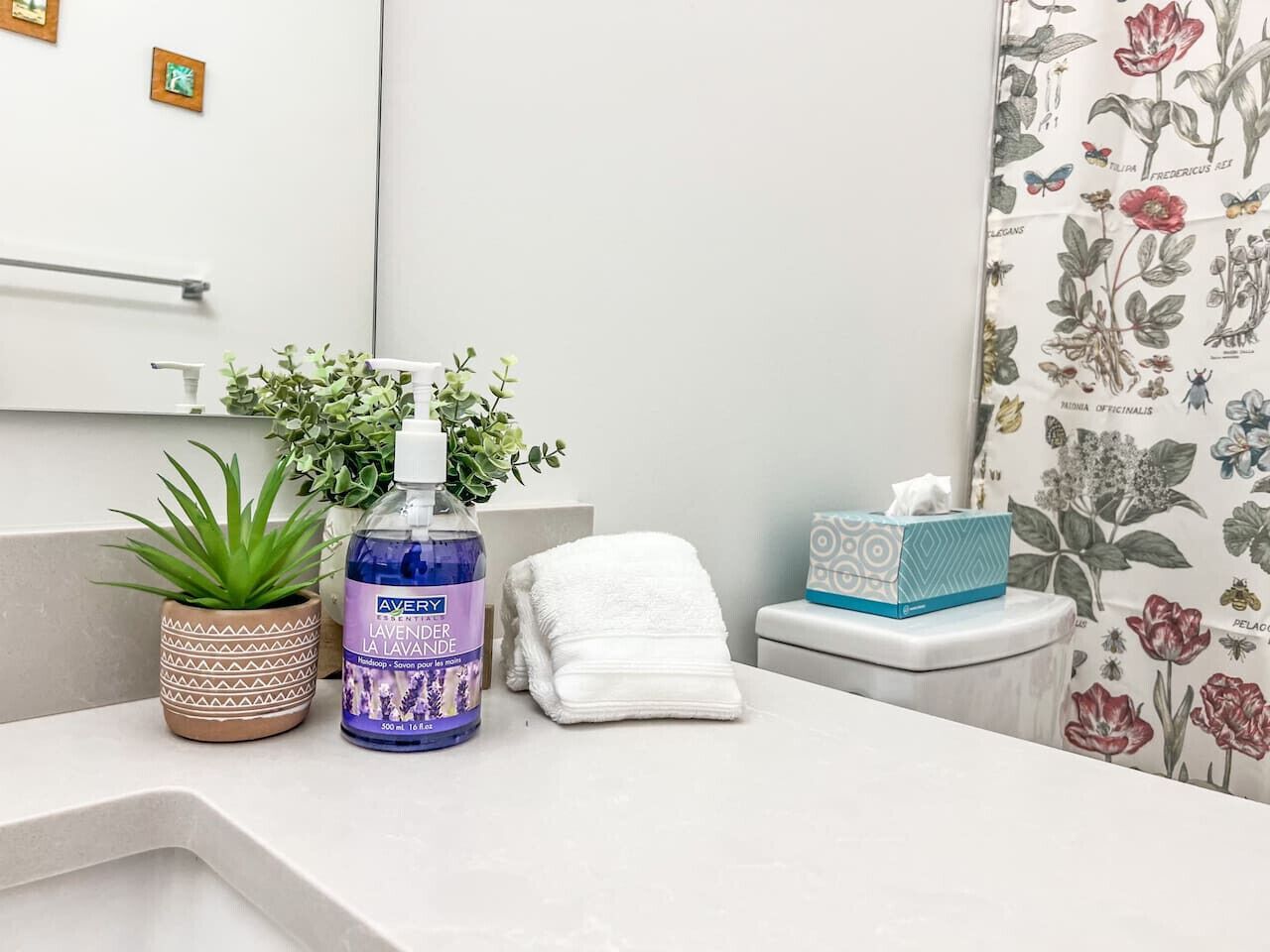 A bathroom with a white sink, a potted plant, a bottle of lavender-scented hand soap, a folded white towel, and a tissue box.