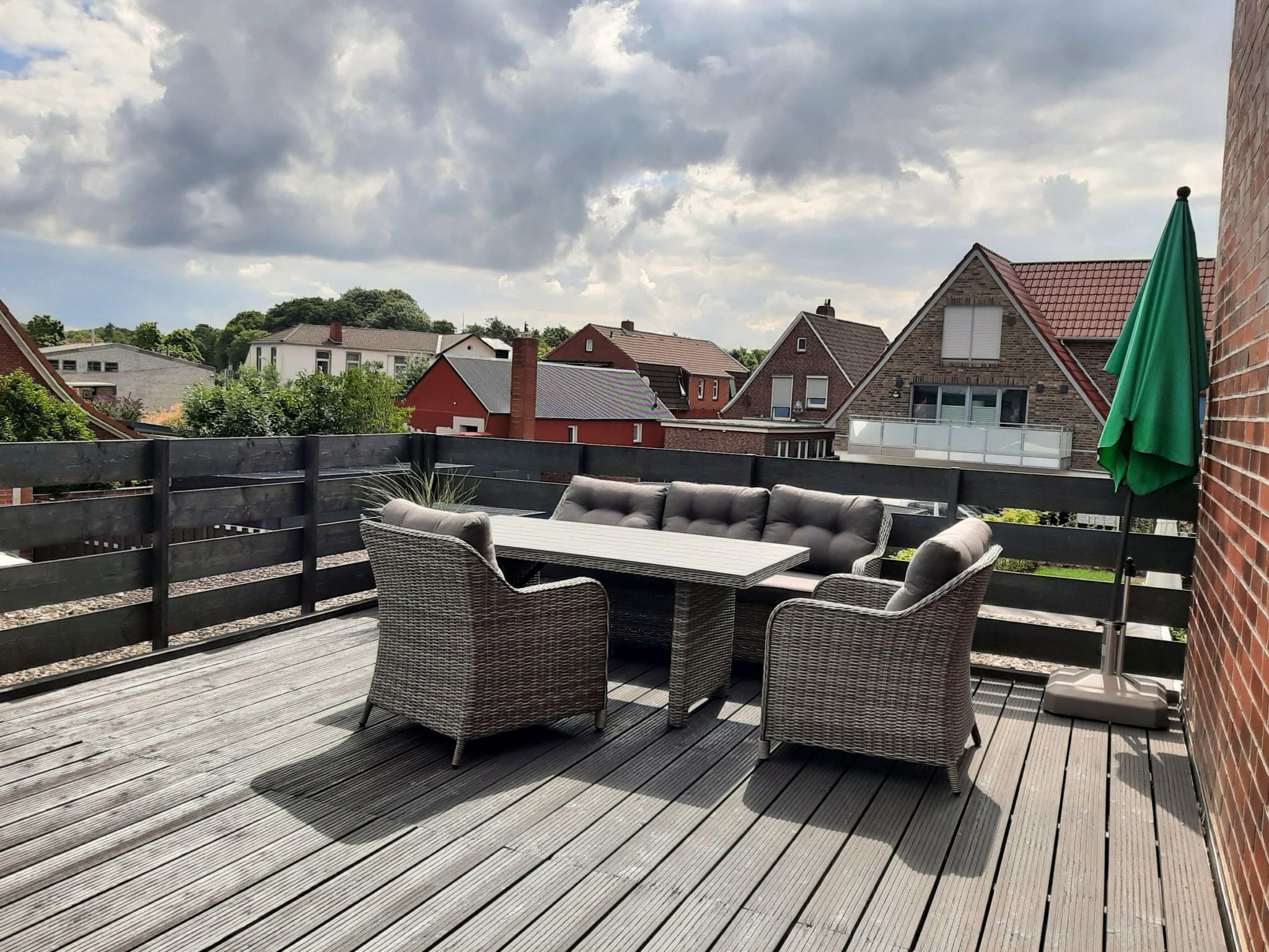 Een houten terras met buitenmeubels, waaronder een tafel en stoelen, en een groene parasol.