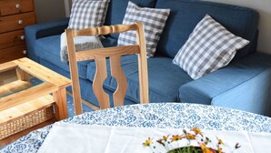 Un canapé bleu agrémenté de coussins à carreaux, une chaise en bois, une petite table en bois avec un plateau en verre et une nappe ornée d’un motif floral bleu.