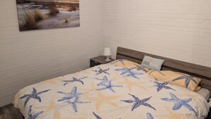 Ein Schlafzimmer mit einem Bett mit einem Meersternmuster, einem Nachttisch, einer Nachttischlampe, einem an der Wand befestigten Bild von einem Sonnenuntergang und einem kleinen Fenster.