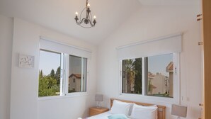 A bedroom with a bed, two bedside tables, a chandelier, and large windows with views of greenery and buildings. - Ferienvilla Protaras 14 by Interhome (Protaras)