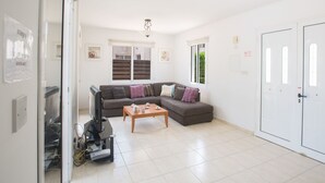 A modern living room with a sectional sofa, a coffee table, and a television. There are windows with blinds, a door, and a ceiling light fixture. - Ferienvilla Protaras 14 by Interhome (Protaras)