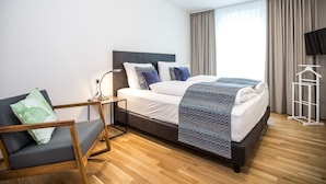 Ein modernes Hotelzimmer mit Bett, Nachttisch, Lampe, Stuhl und einem an der Wand montierten Fernseher.