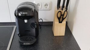 En köksbänk med en Bosch-kaffemaskin och en TASSIMO-högtalare, ett knivblock i trä samt en låda med prydligt ordnade bestick och köksredskap.